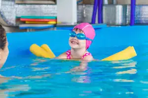 A little girl swims in a pool with a pnik cap, blue goggles, and yellow pool noodles.
